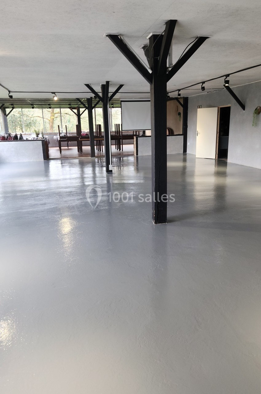 Salle spacieuse avec sol en béton gris brillant, poutres noires et grandes fenêtres laissant entrer la lumière naturelle.