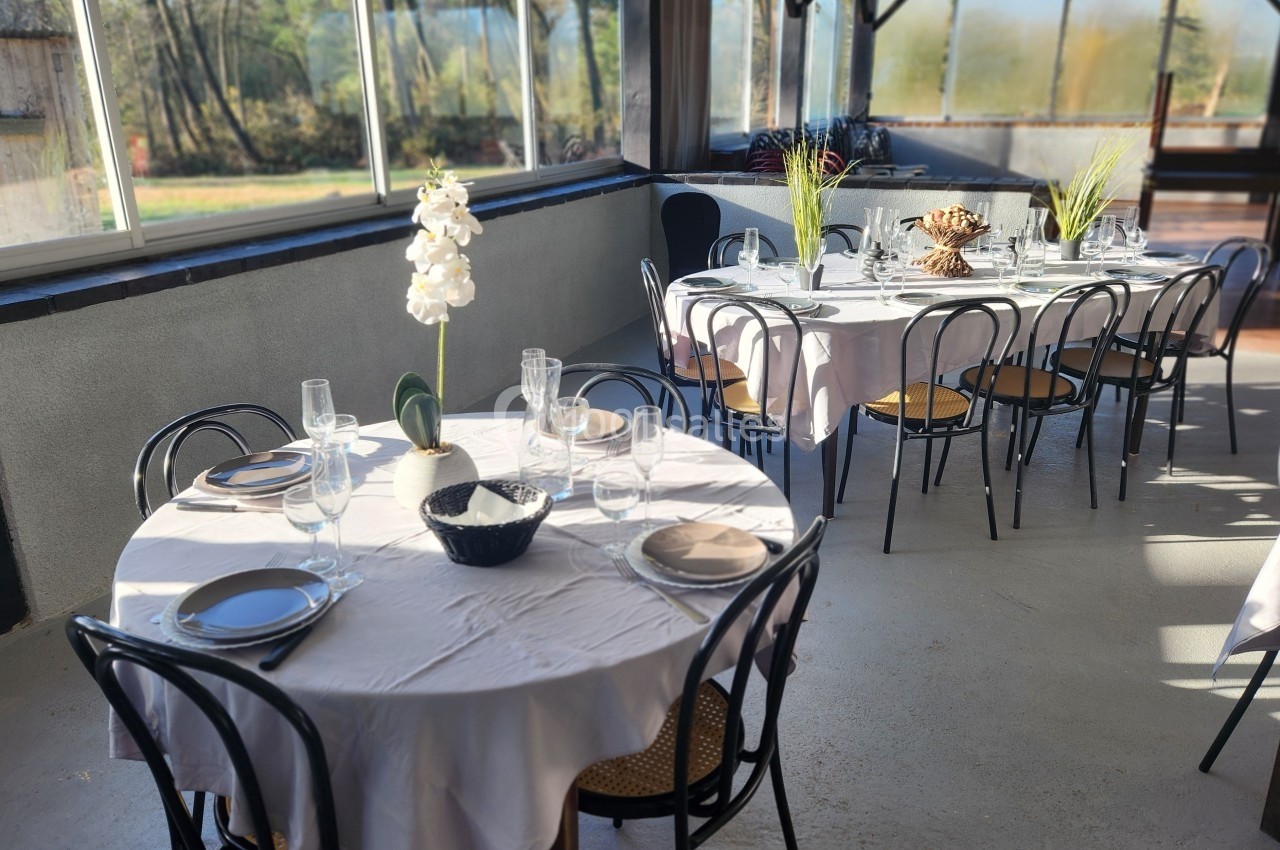 Salle lumineuse avec tables rondes dressées, nappes blanches, chaises noires et vue sur un extérieur arboré.