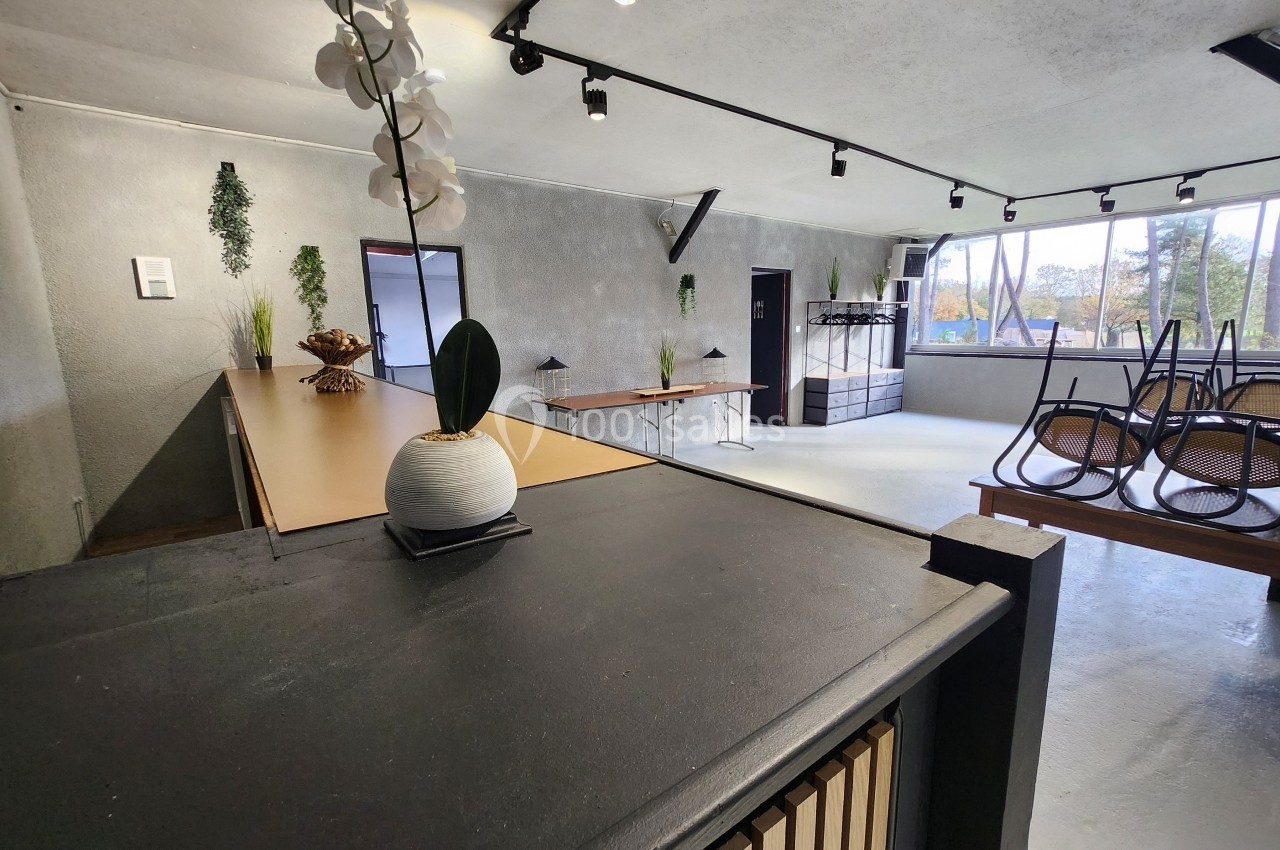 Espace intérieur lumineux avec tables empilées, plantes décoratives et grandes fenêtres laissant entrer la lumière naturelle.