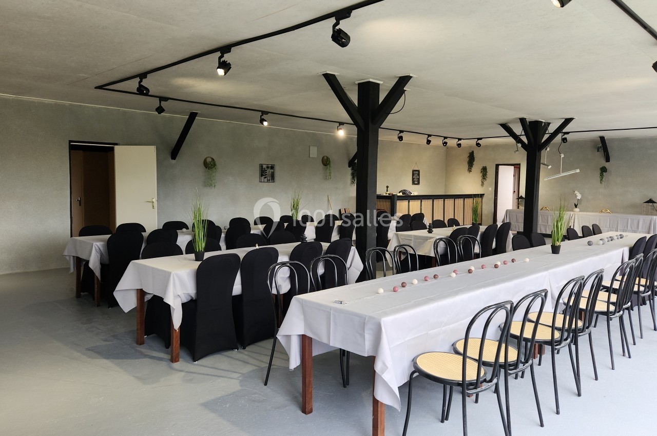 Salle de réception avec tables dressées, nappes blanches, chaises noires et décoration sobre, éclairée par des spots au…