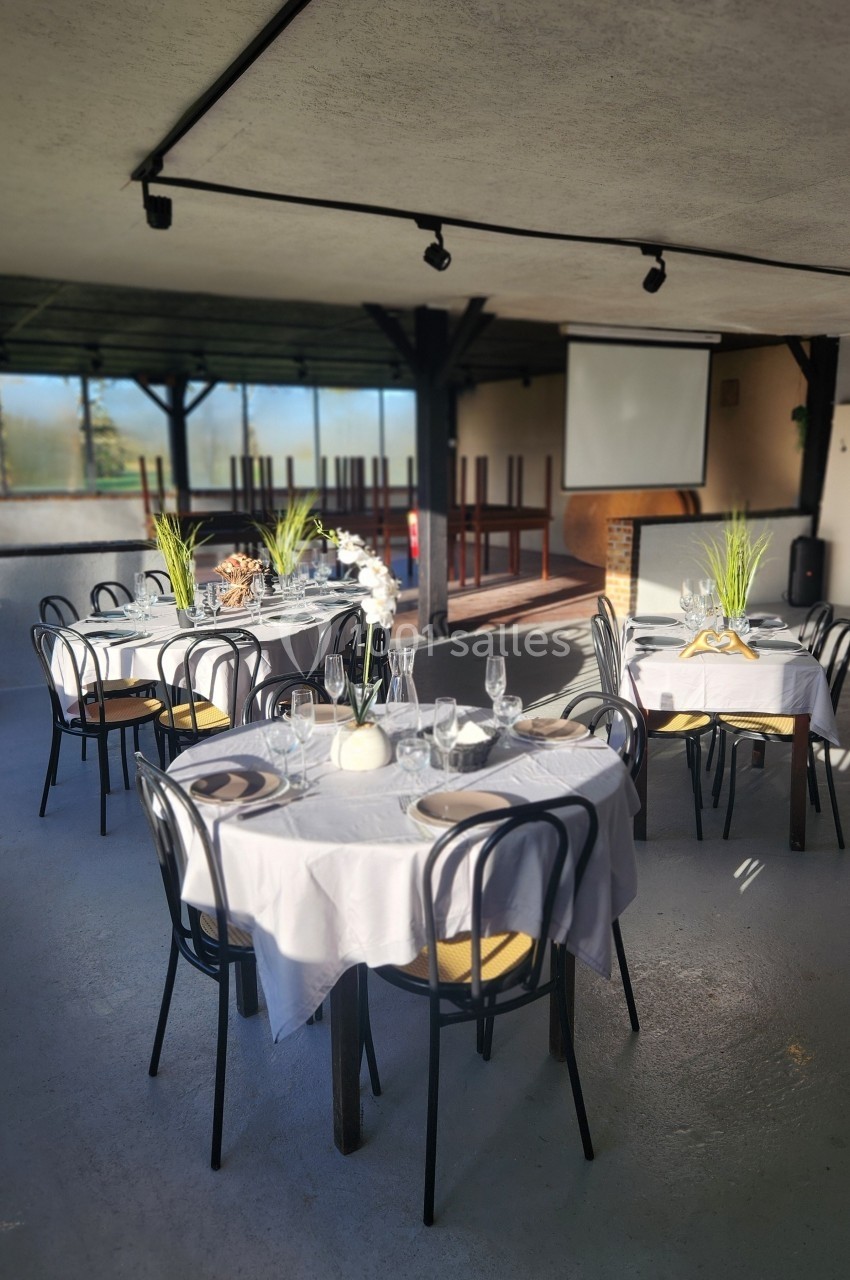 Salle lumineuse avec tables rondes dressées pour un repas, nappes blanches, verres et décorations végétales.
