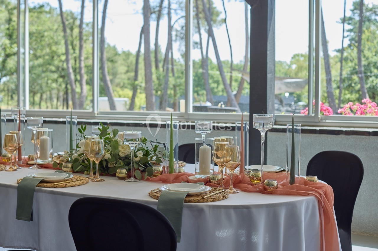 Table élégamment dressée avec nappes blanches, décor floral, bougies et vaisselle dorée, dans une salle lumineuse avec vue…