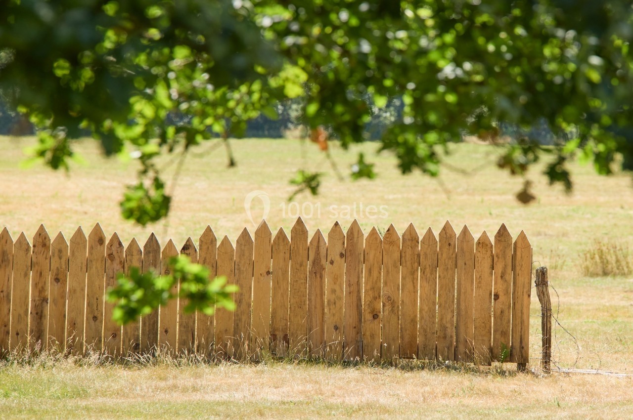 Clôture en bois dans un champ ensoleillé, partiellement ombragée par des branches d'arbres au premier plan.