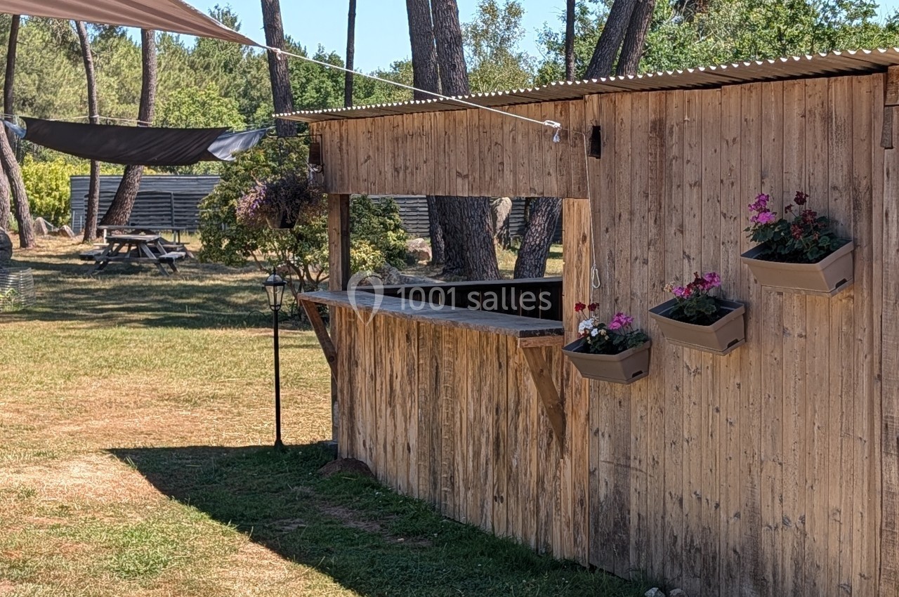 Kiosque en bois avec jardinières fleuries, situé dans un espace extérieur arboré et ensoleillé.