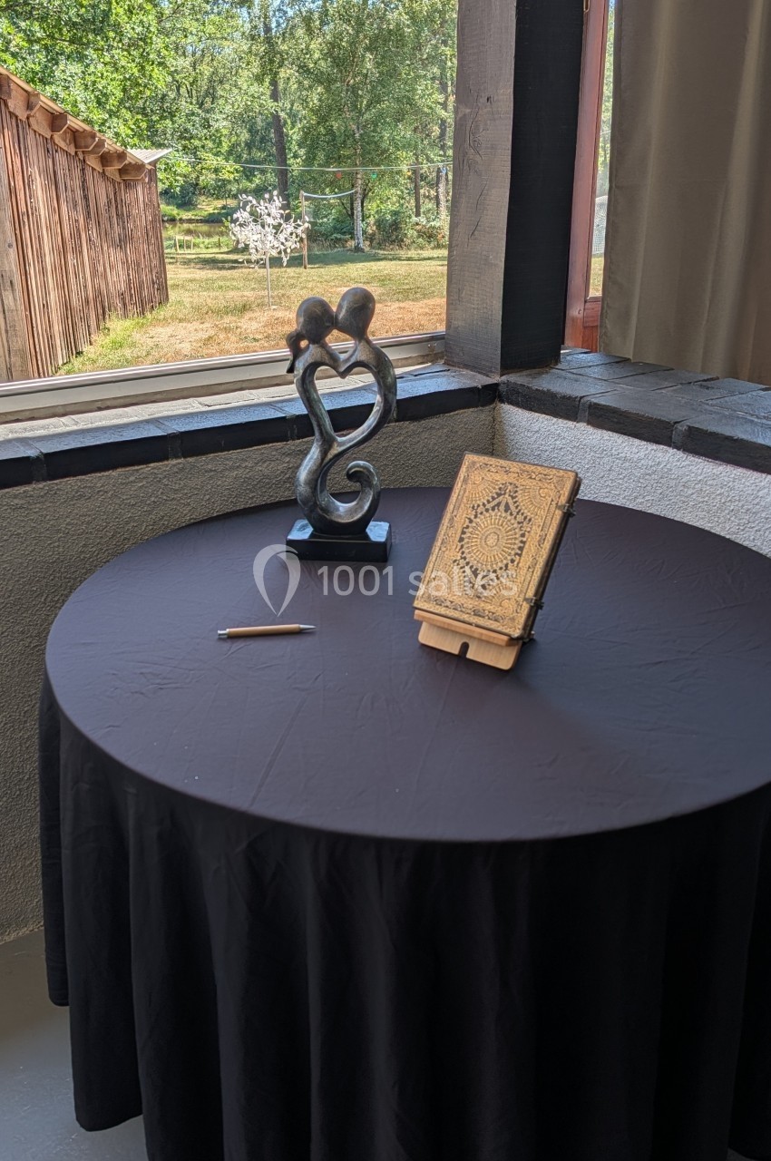 Table ronde avec nappe noire, statue en métal représentant un couple, livre ancien ouvert et stylo, près d'une fenêtre.