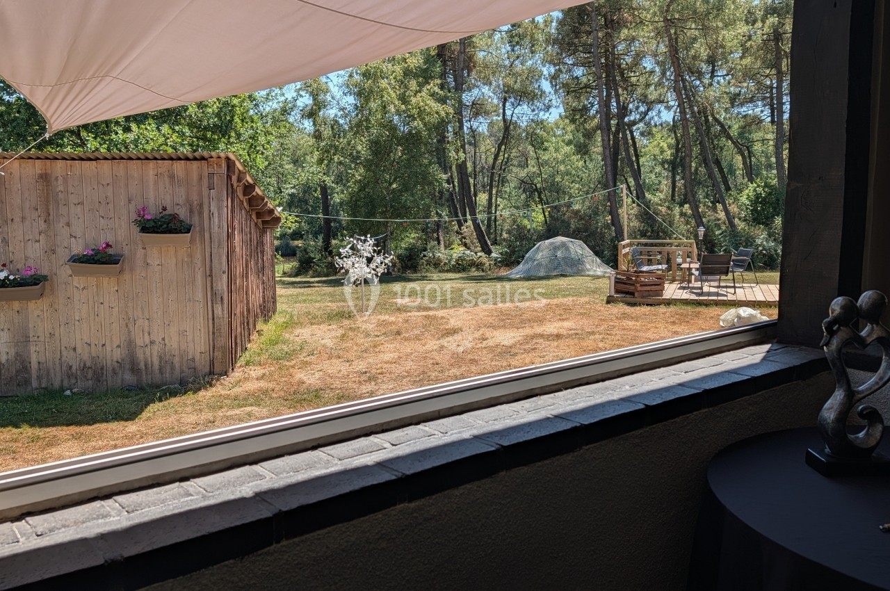 Vue d'un jardin avec une cabane en bois, des fleurs suspendues, une pelouse sèche et des arbres en arrière-plan.