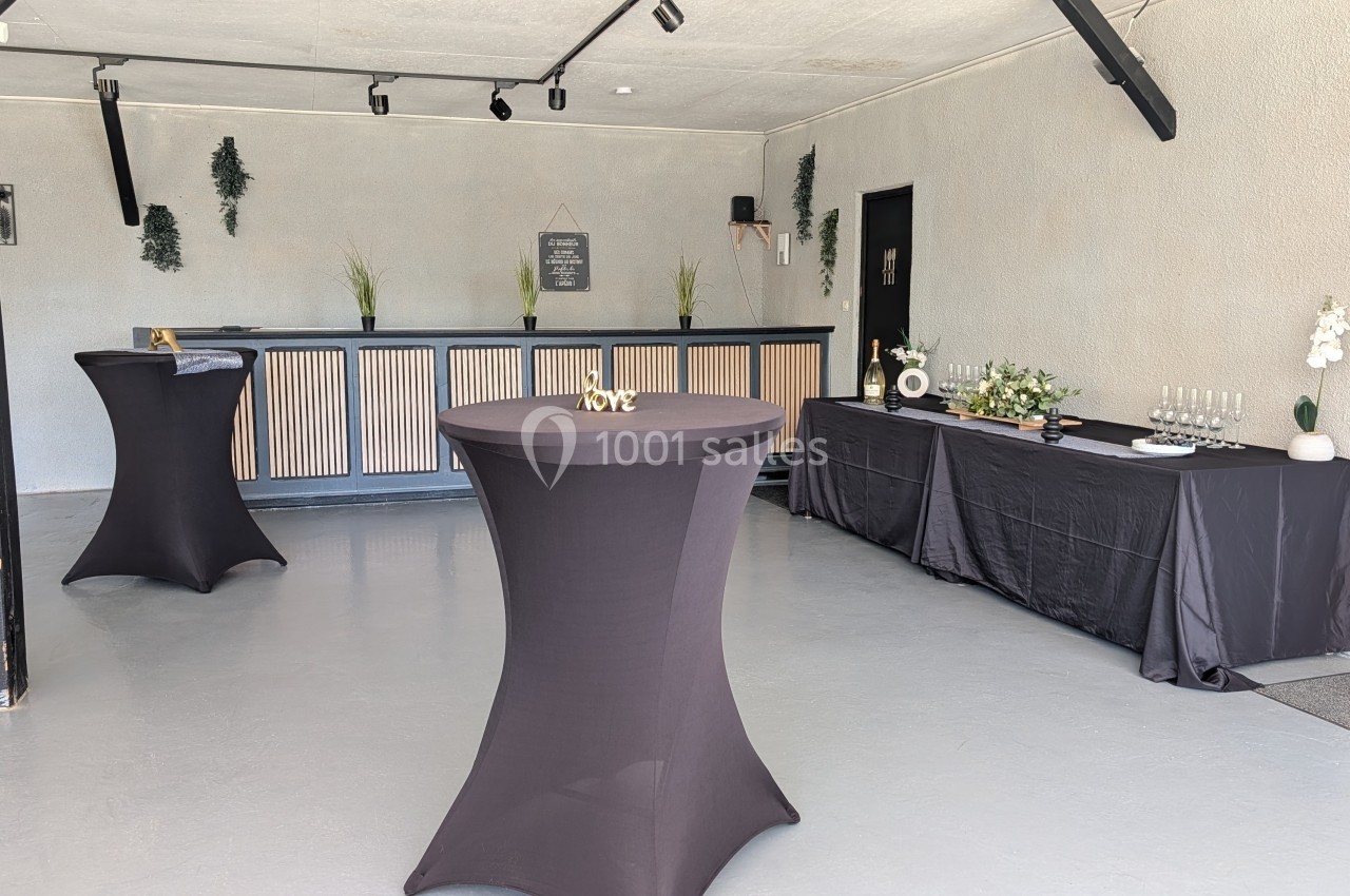 Salle aménagée avec des tables hautes noires, un comptoir en bois et une table de service décorée pour un événement.
