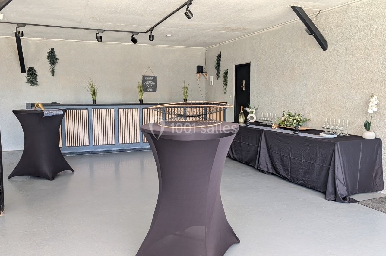 Salle sobre avec comptoir en bois, tables hautes drapées de noir et buffet décoré de fleurs et verres.