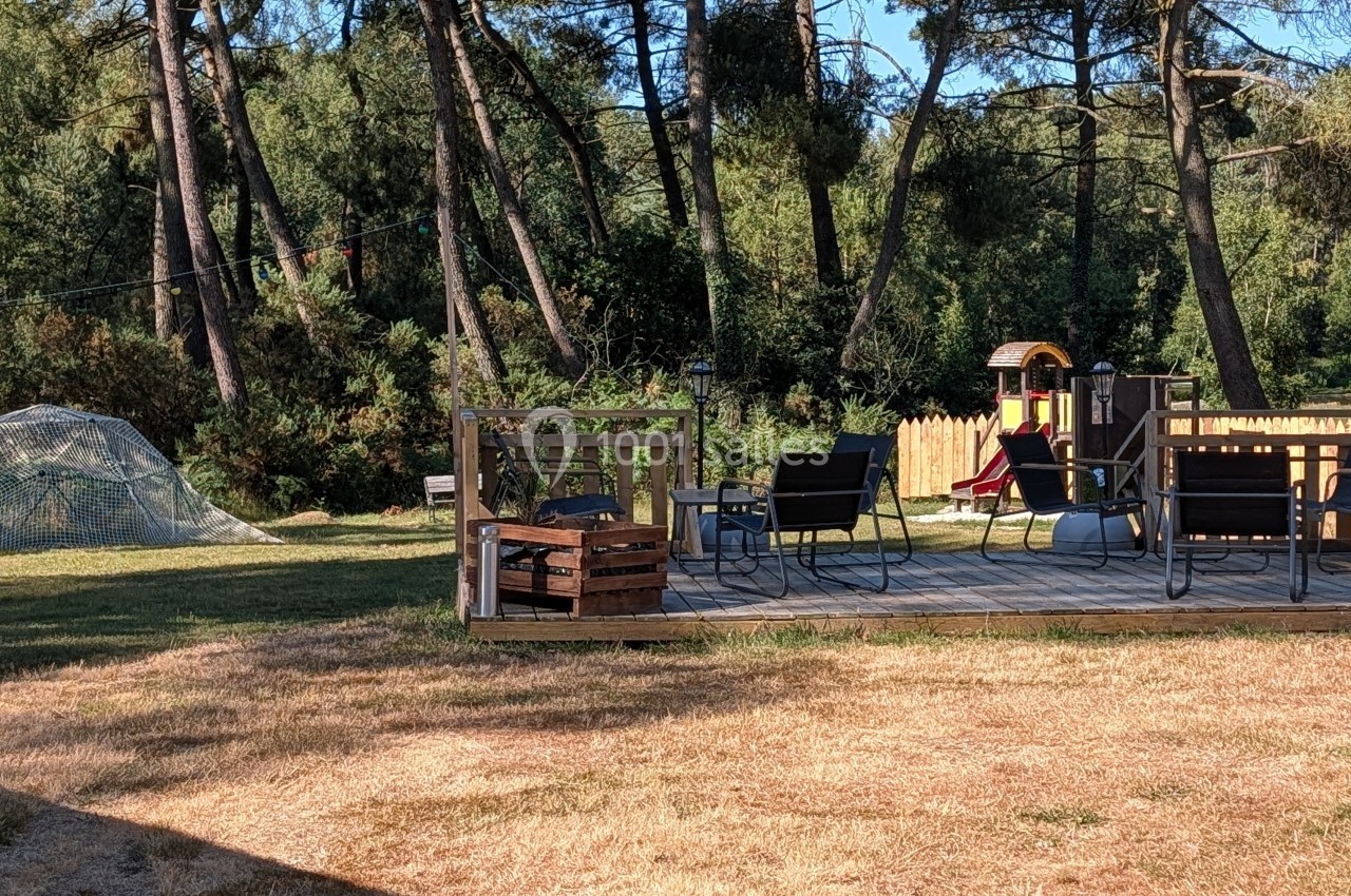 Terrasse en bois avec des chaises et un brasero, entourée d'arbres, avec une aire de jeux et une structure en filet à…