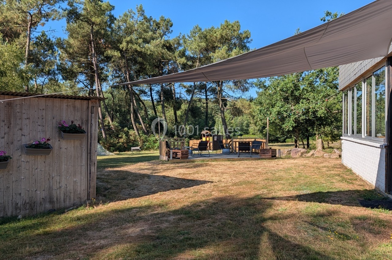 Espace extérieur avec pelouse, terrasse en bois, tables, arbres en arrière-plan et voile d'ombrage tendue.
