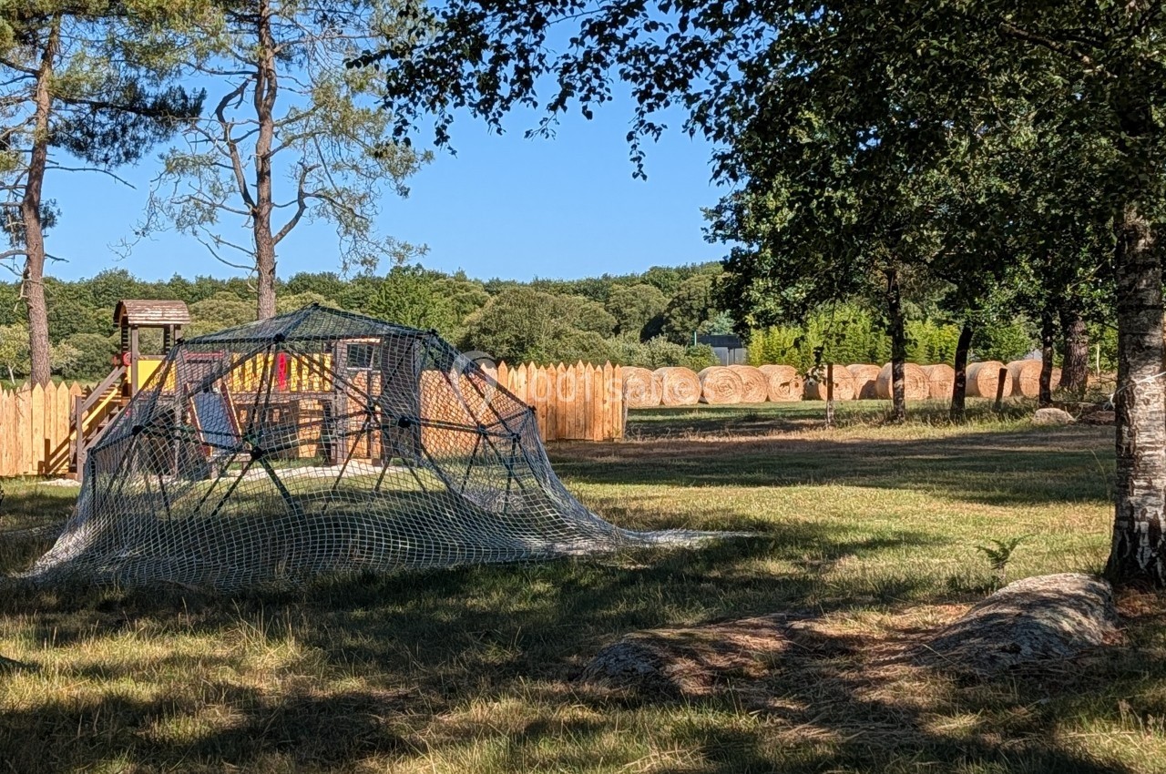 Structure de jeu en bois et filet dans un espace extérieur avec arbres, clôture en bois et bottes de foin en arrière-plan.