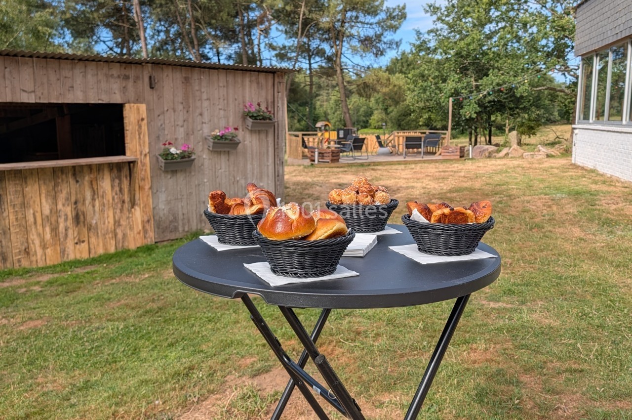 Pains et viennoiseries disposés dans des paniers noirs sur une table ronde en extérieur, près d'un bâtiment en bois.