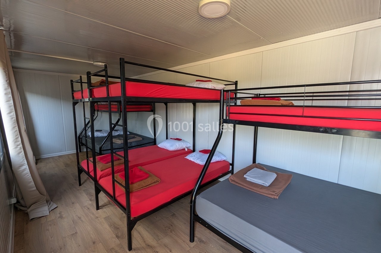 Chambre simple avec lits superposés et un lit simple, draps rouges et gris, sol en bois et murs clairs.