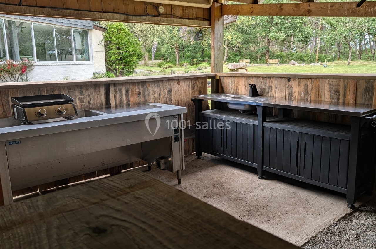 Cuisine extérieure couverte en bois avec plan de travail en inox, barbecue, rangements noirs et vue sur un jardin boisé.