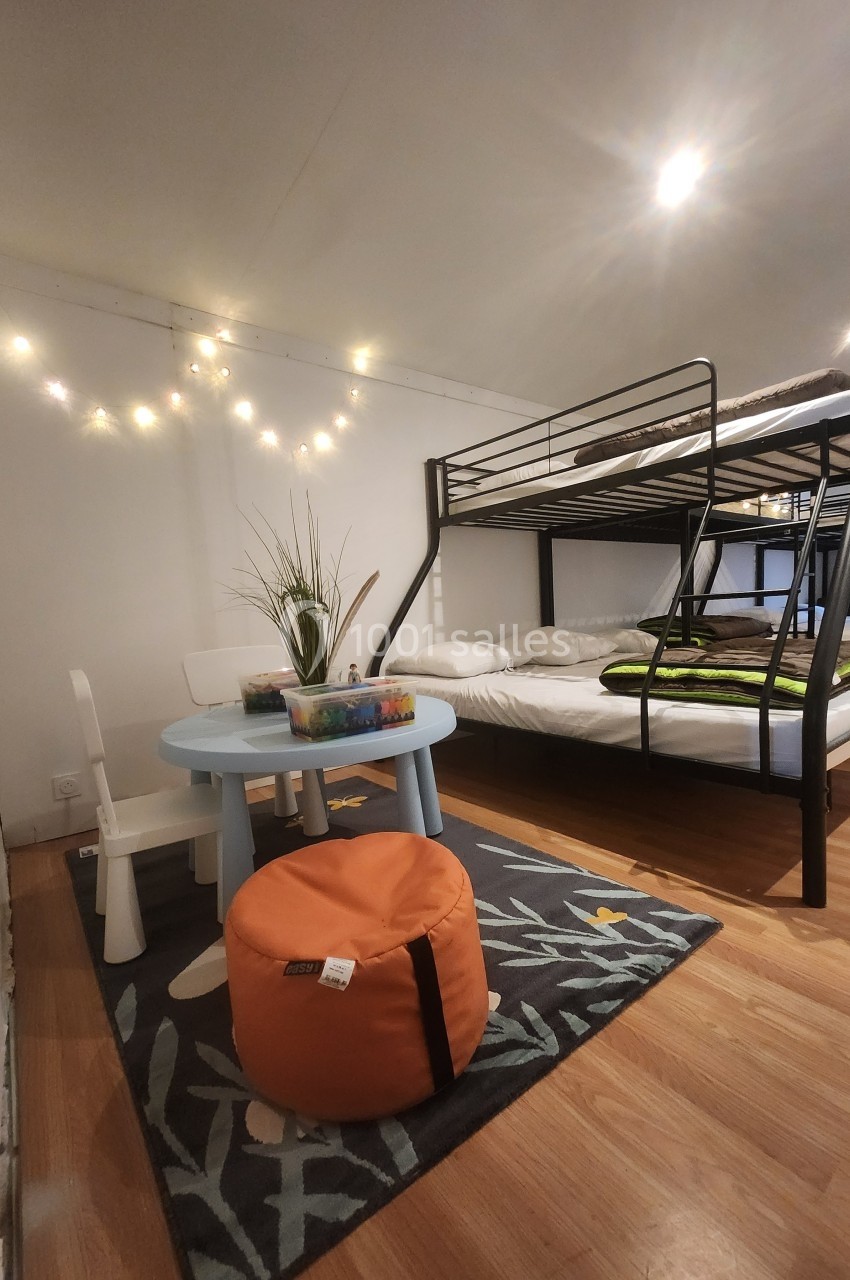 Chambre avec lits superposés, table basse, chaise enfant, pouf orange et guirlande lumineuse sur mur blanc.