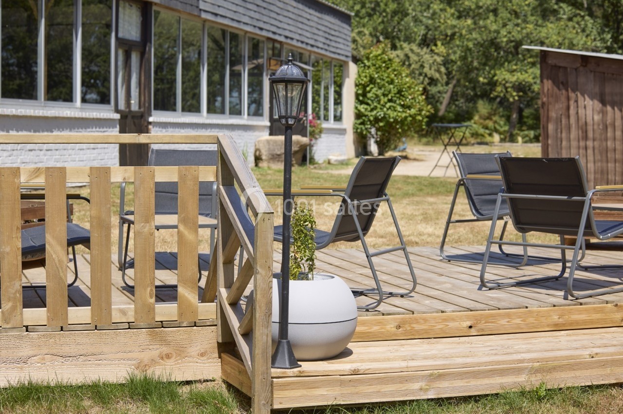 Terrasse en bois avec chaises et table, entourée de verdure, près d'un bâtiment vitré et d'un lampadaire.