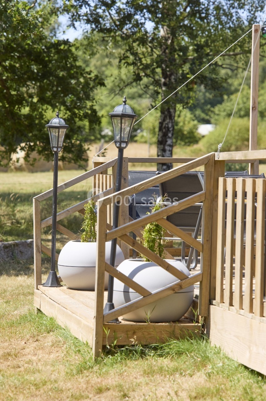 Terrasse en bois avec balustrade, pots de fleurs blancs, lampadaires noirs et vue sur un jardin arboré.