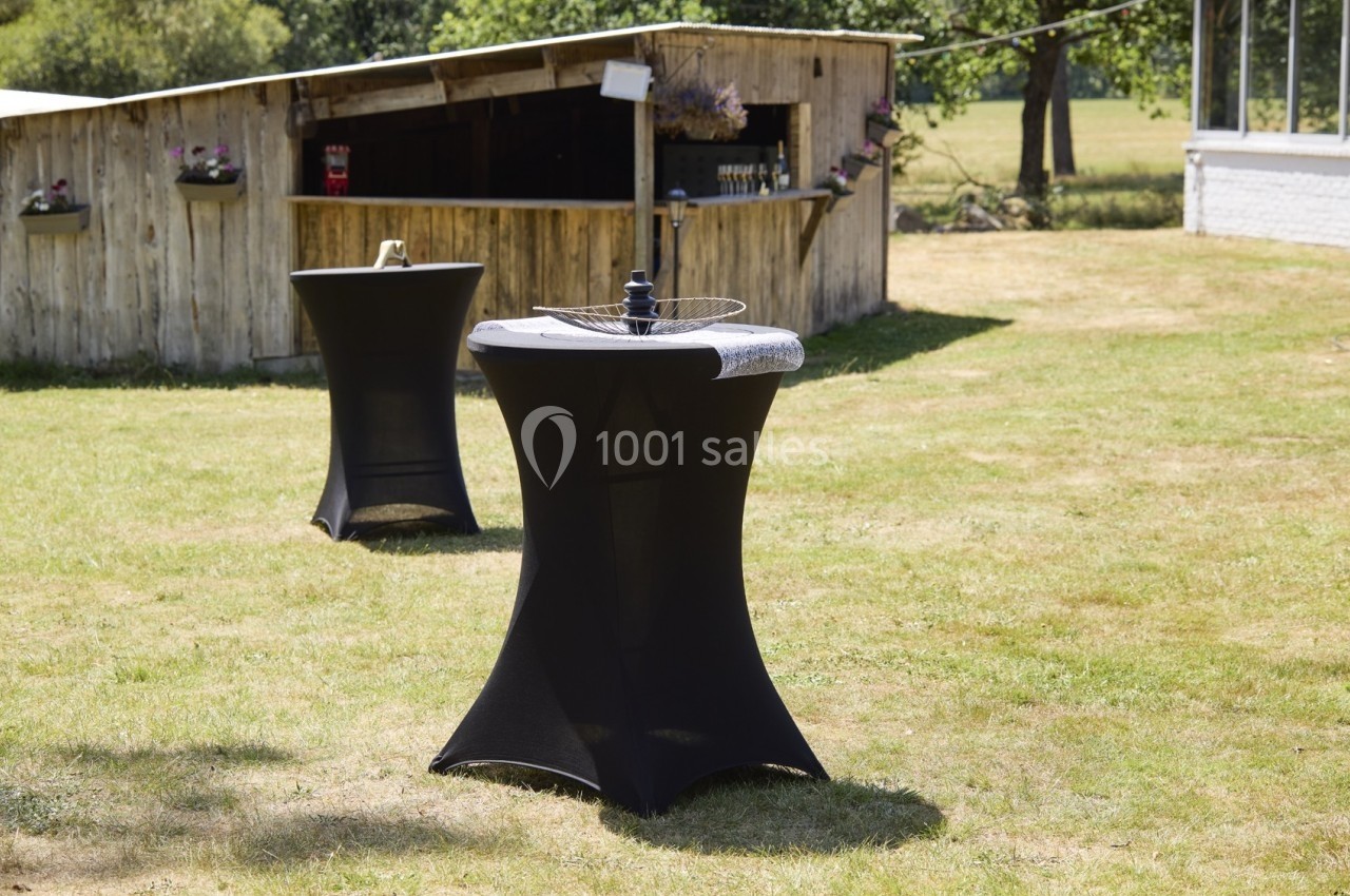 Table haute noire avec housse, posée sur une pelouse près d'un bar extérieur en bois.