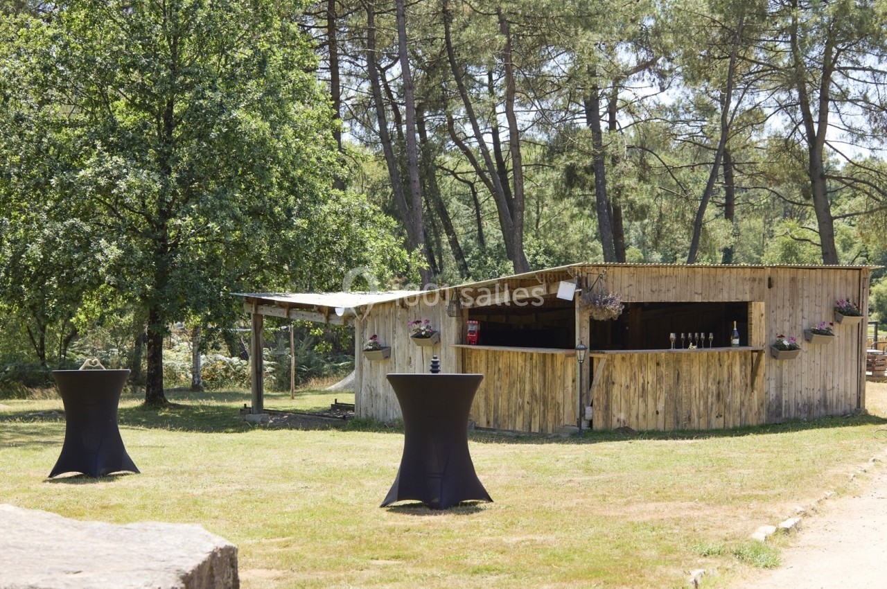 Bar en bois en extérieur entouré d'arbres, avec deux tables hautes noires sur une pelouse.
