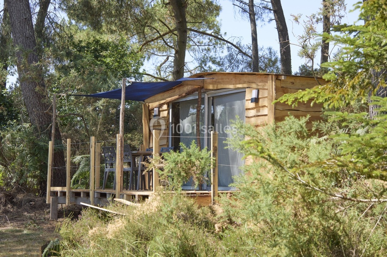 Cabane en bois avec terrasse ombragée, située dans une forêt entourée de végétation dense.