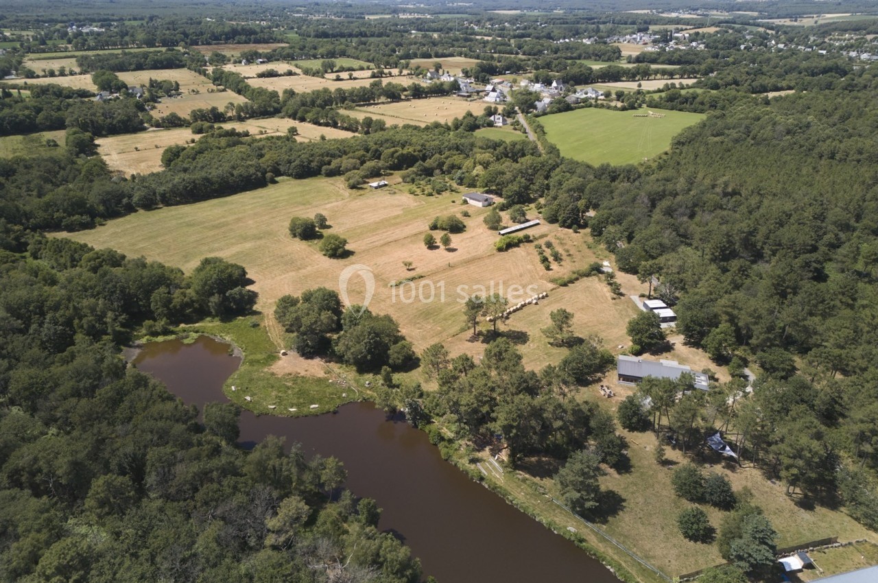 Vue aérienne d'une campagne avec champs, forêt, étang et quelques bâtiments dispersés sous un ciel dégagé.