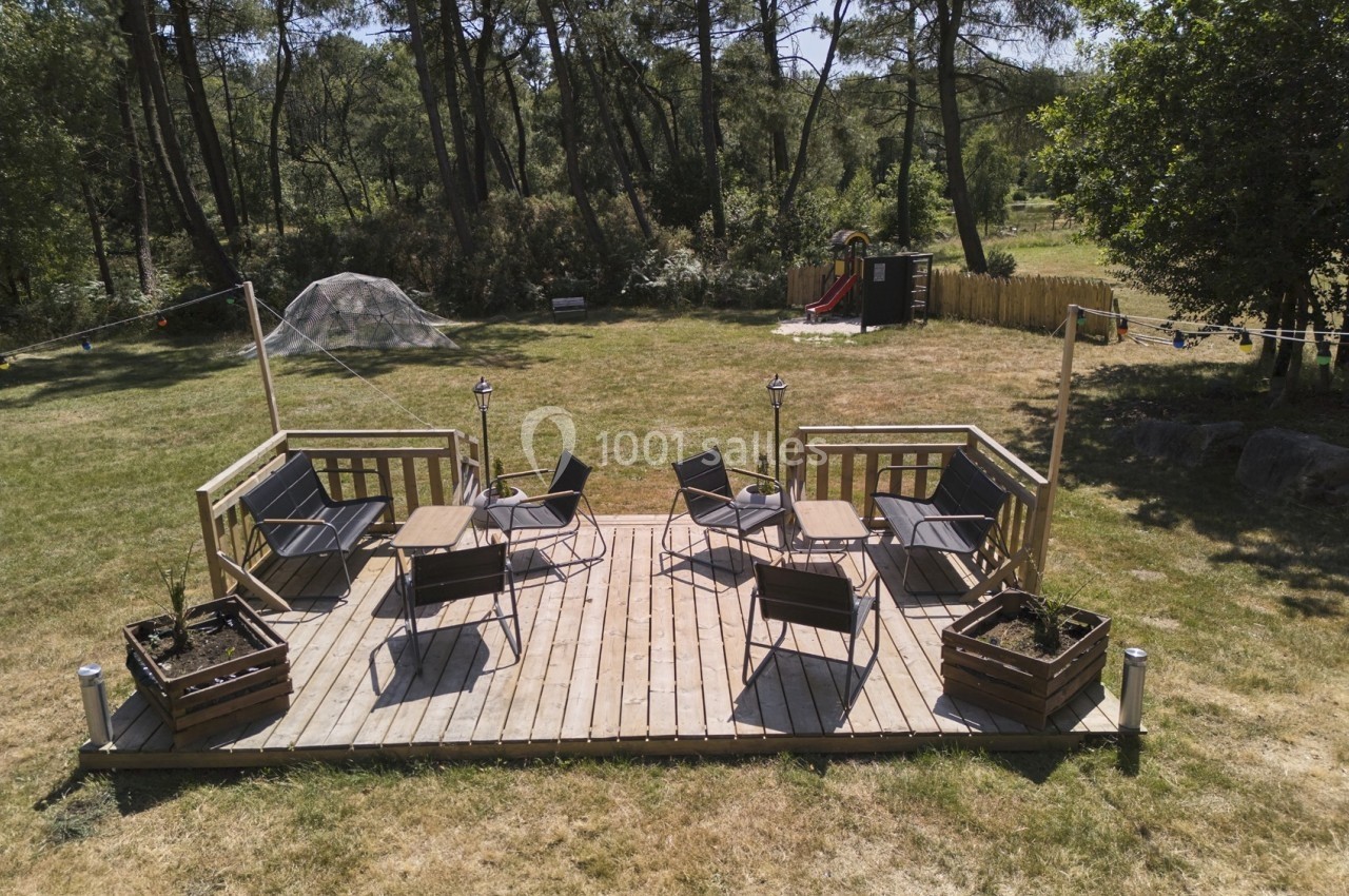 Terrasse en bois avec chaises et tables, entourée de pelouse, arbres et jeux pour enfants en arrière-plan.