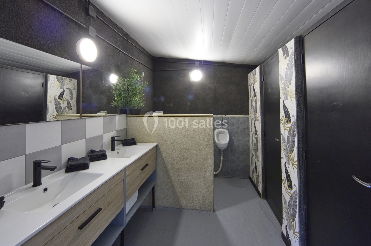 Salle de bain moderne avec deux lavabos, un urinoir, des murs sombres et une décoration végétale.