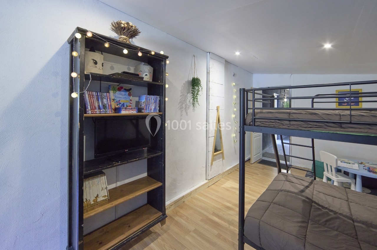 Étagère avec livres, guirlande lumineuse et télévision, lit superposé et table enfant dans une pièce lumineuse.