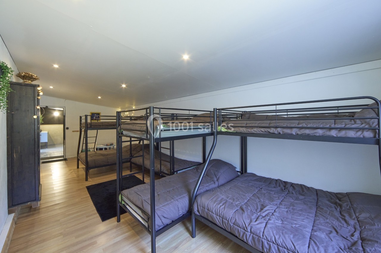 Dortoir avec lits superposés en métal, matelas bruns et sol en parquet, menant à une salle de bain au fond.