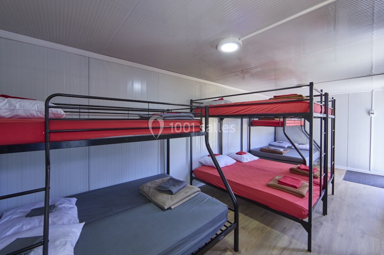 Dortoir avec lits superposés en métal, matelas rouges et gris, draps pliés, dans une pièce lumineuse au sol en bois.