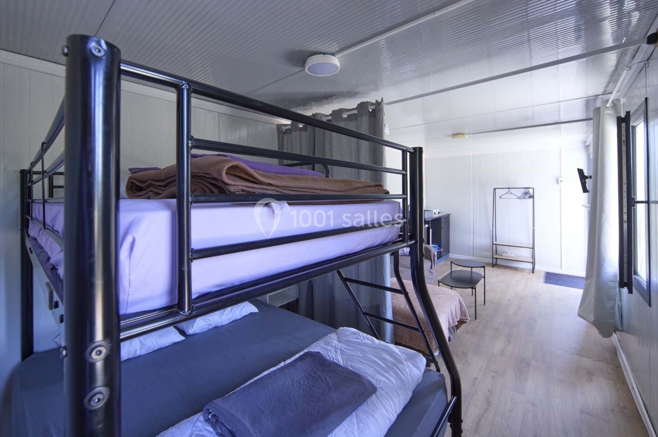 Chambre simple avec lits superposés, draps pliés, sol en bois clair et lumière naturelle entrant par une fenêtre.
