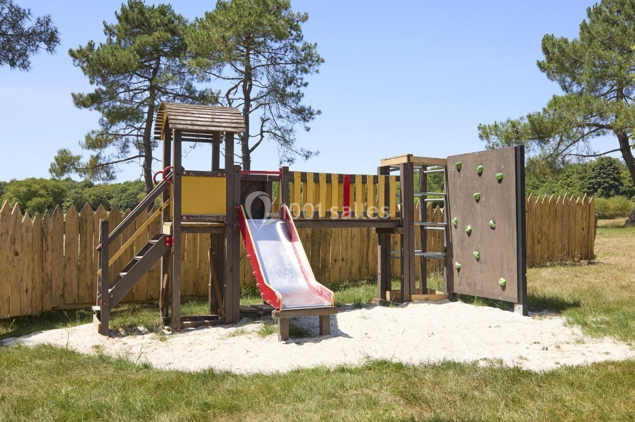Structure de jeu en bois avec toboggan rouge, mur d'escalade et échelle, située sur un sol sablonneux en extérieur.