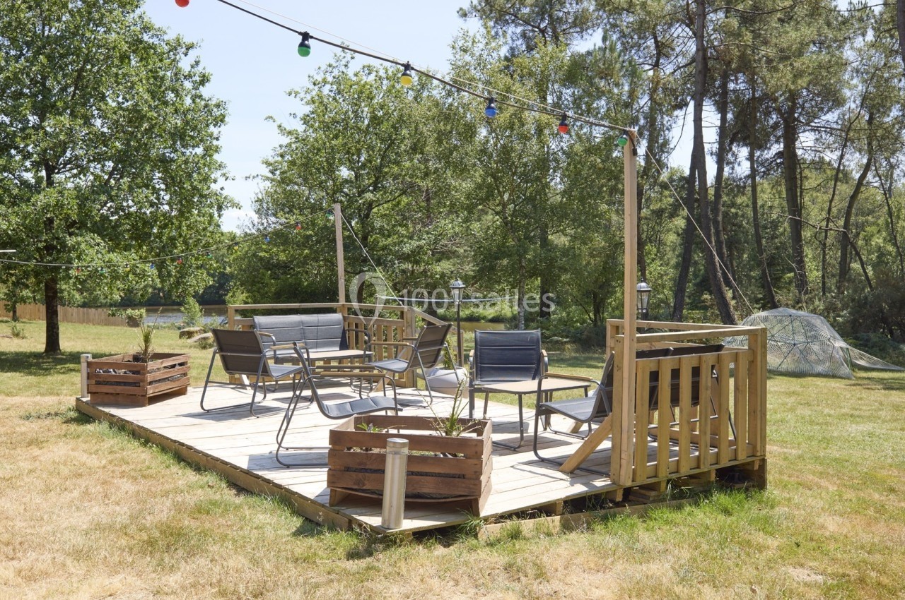 Terrasse en bois avec mobilier de jardin, entourée de verdure et éclairée par des guirlandes lumineuses colorées.