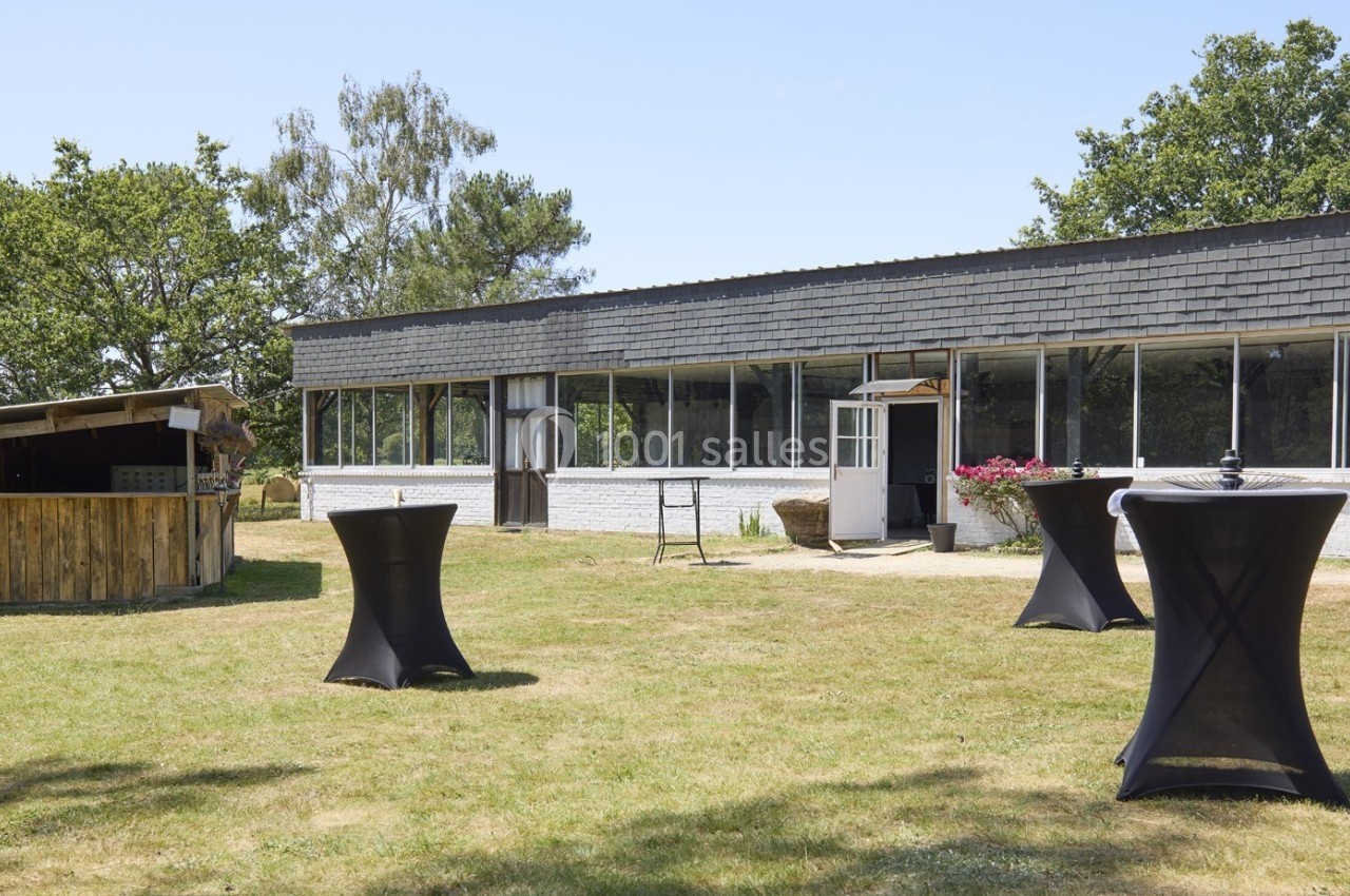 Espace extérieur avec tables hautes noires sur une pelouse, devant un bâtiment vitré et un comptoir en bois.