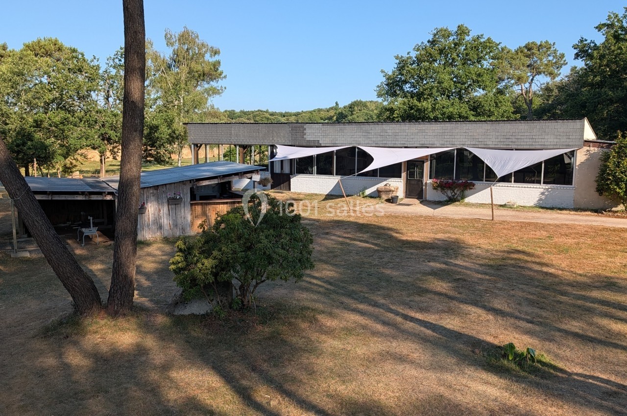 Bâtiment moderne avec grandes baies vitrées et voiles d'ombrage, entouré d'arbres et d'une pelouse sèche.