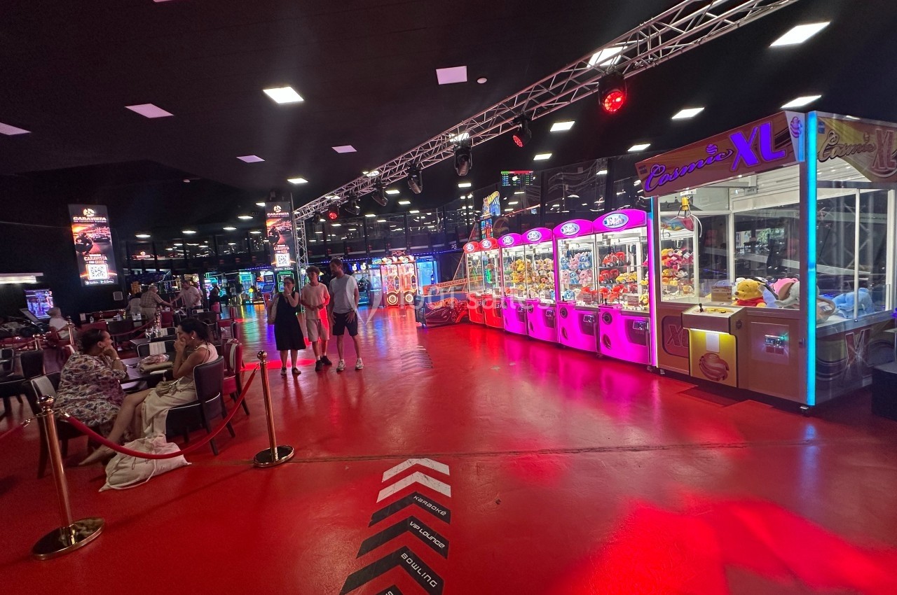 Salle de jeux avec sol rouge, machines à pinces lumineuses et visiteurs explorant l'espace.