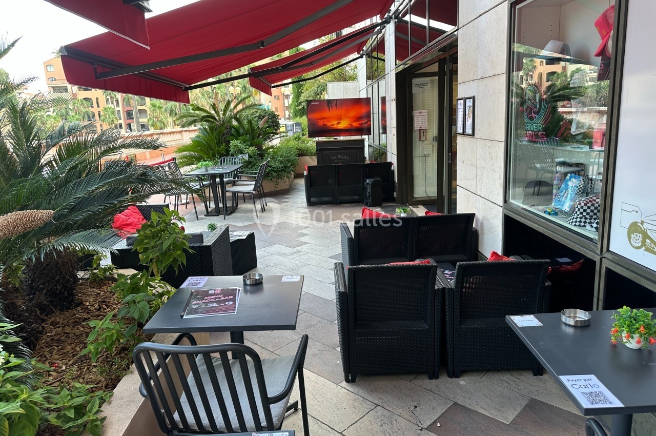 Terrasse extérieure d'un café avec tables, chaises, canapés et écran, entourée de plantes et protégée par des stores rouges.