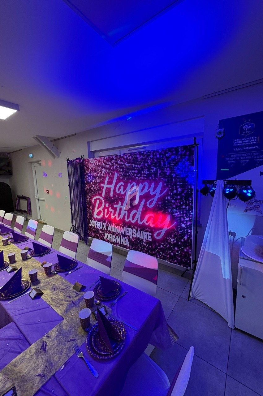 Salle décorée pour un anniversaire avec une longue table dressée en violet et un panneau lumineux ’Happy Birthday’.