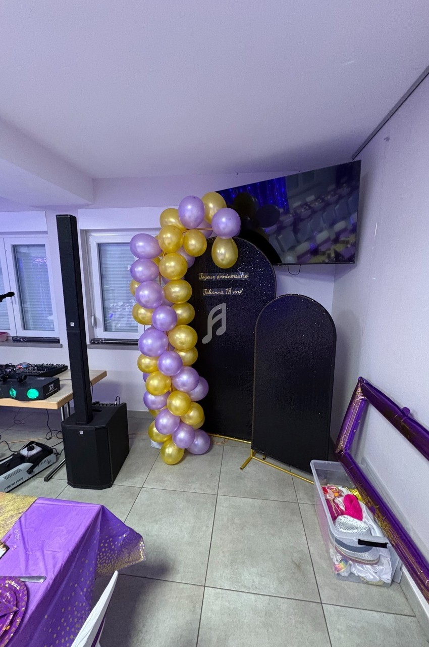 Guirlande de ballons violets et dorés décorant un coin de salle avec écran, panneaux noirs et matériel audio.