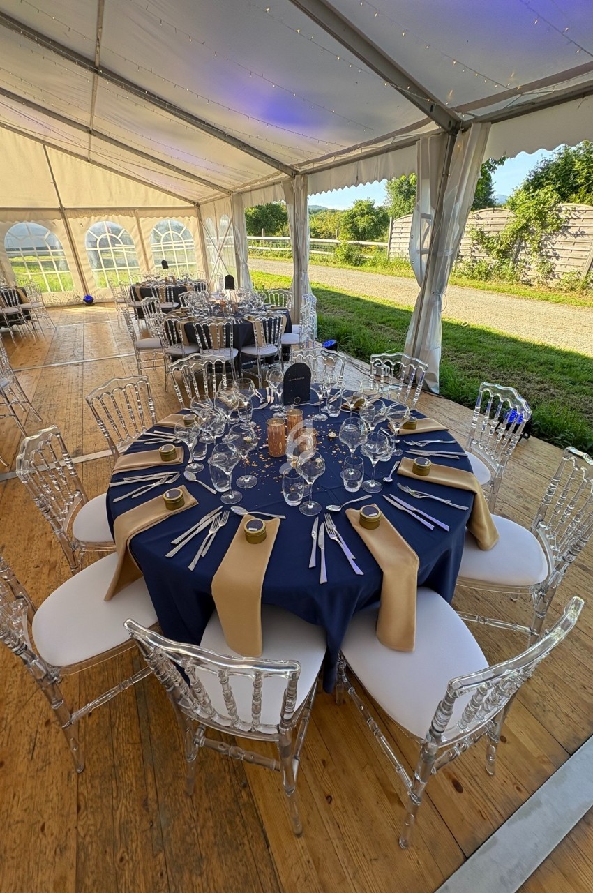 Table dressée avec une nappe bleue, serviettes dorées, verres et couverts, sous une tente lumineuse avec chaises…