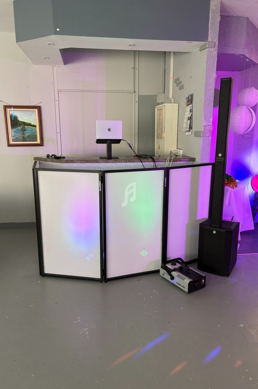 Pupitre de DJ avec éclairage coloré, haut-parleur vertical et matériel audio dans une salle sobrement décorée.
