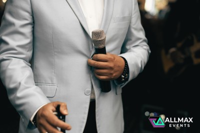 Un homme en costume clair tenant un microphone, prêt à parler lors d'un événement en intérieur.