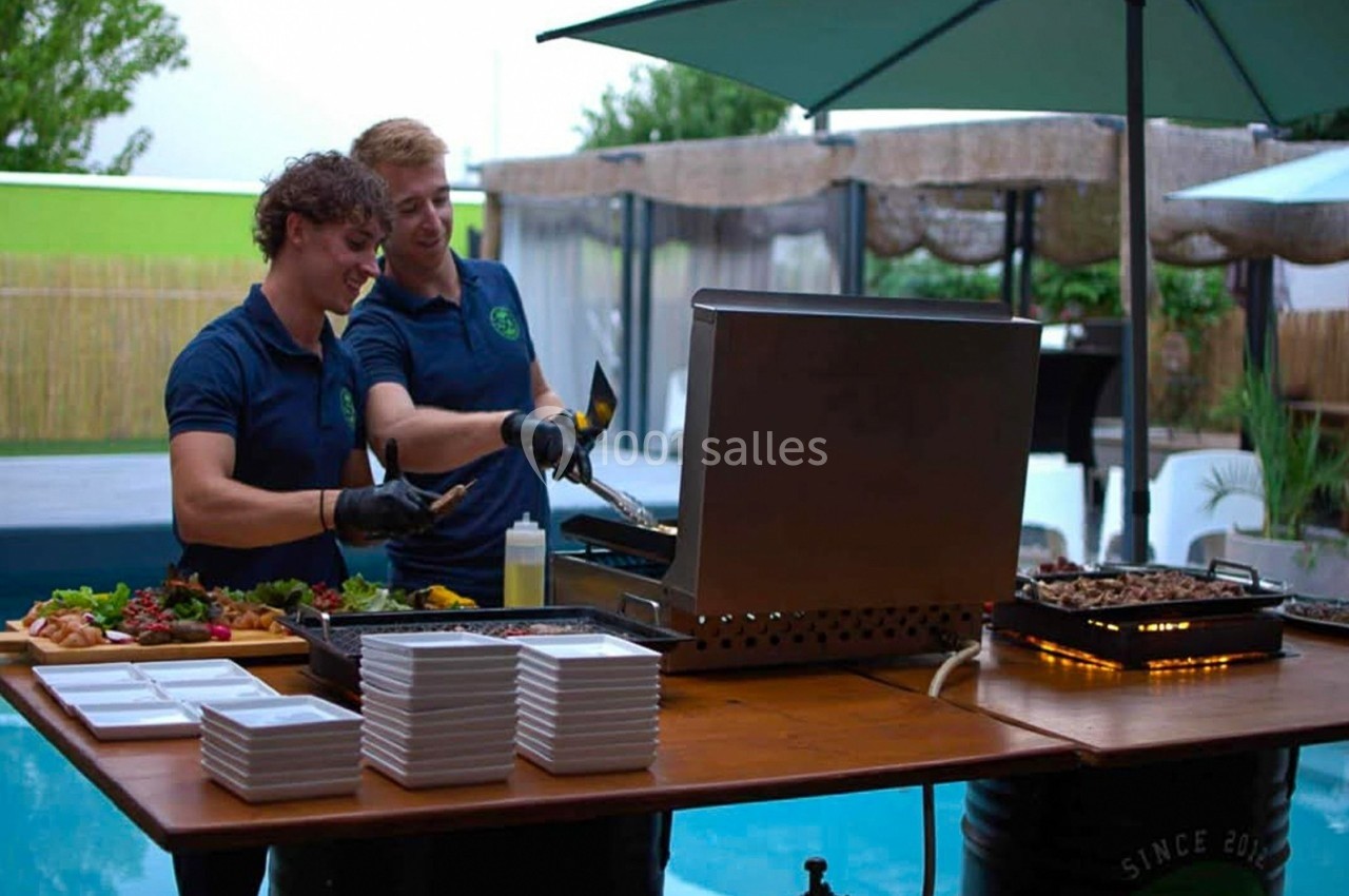 Deux cuisiniers préparent des plats sur une plancha près d'une piscine, entourés de plateaux et d'ustensiles.