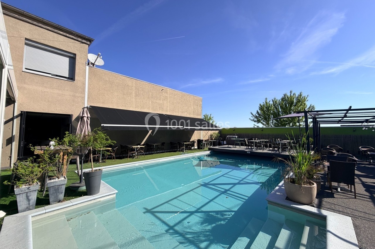 Piscine extérieure entourée de plantes, avec terrasse ombragée et mobilier de jardin sous un ciel dégagé.