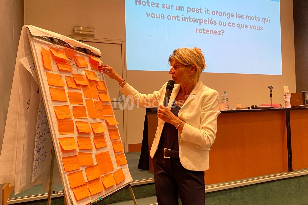 Une femme présente des idées écrites sur des post-it orange collés sur un tableau lors d'une conférence.