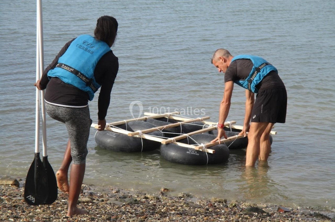 Deux personnes en gilets de sauvetage ajustent un radeau fait de pneus et de bois au bord de l'eau.