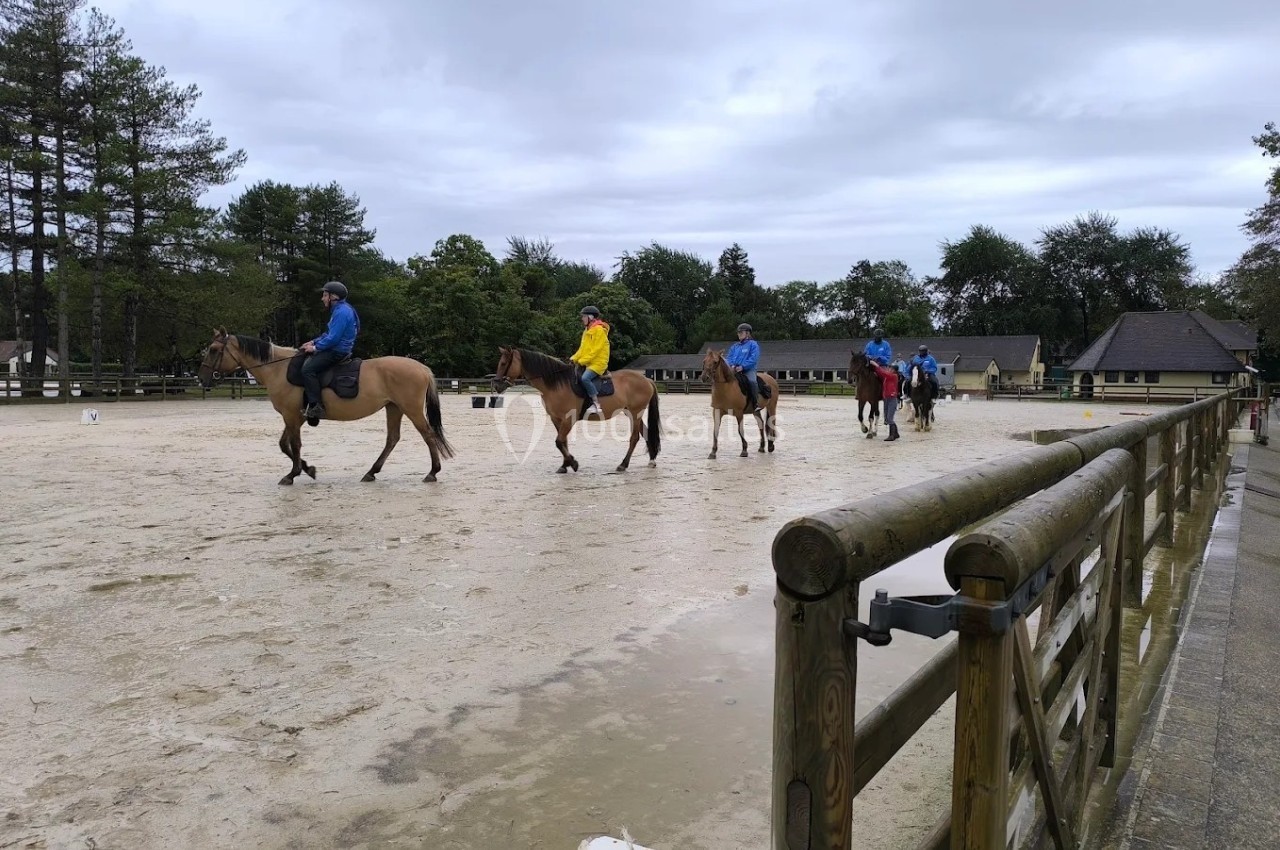 Des cavaliers en file montent des chevaux sur une grande carrière extérieure par temps nuageux.