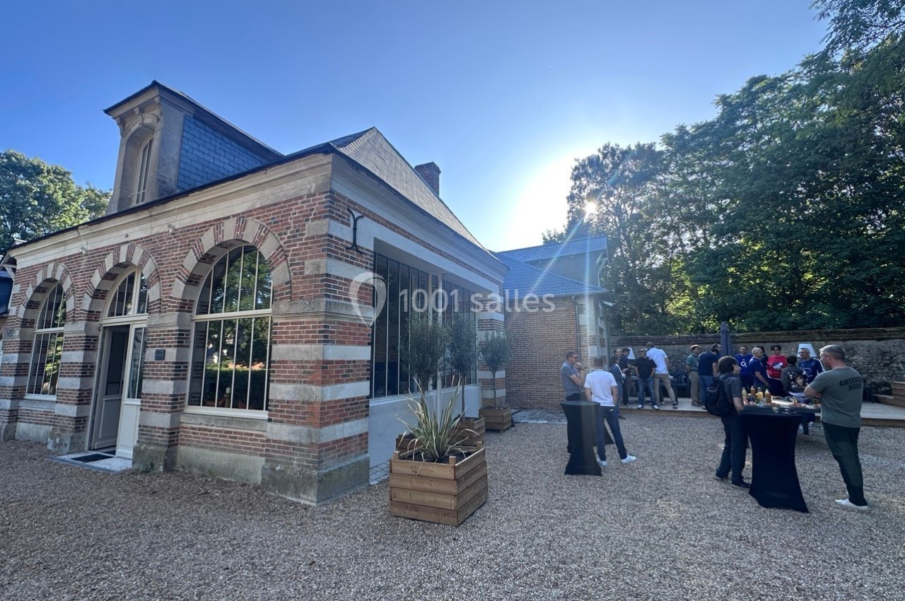 Bâtiment en briques avec grandes fenêtres, terrasse en gravier, personnes discutant autour de tables hautes.