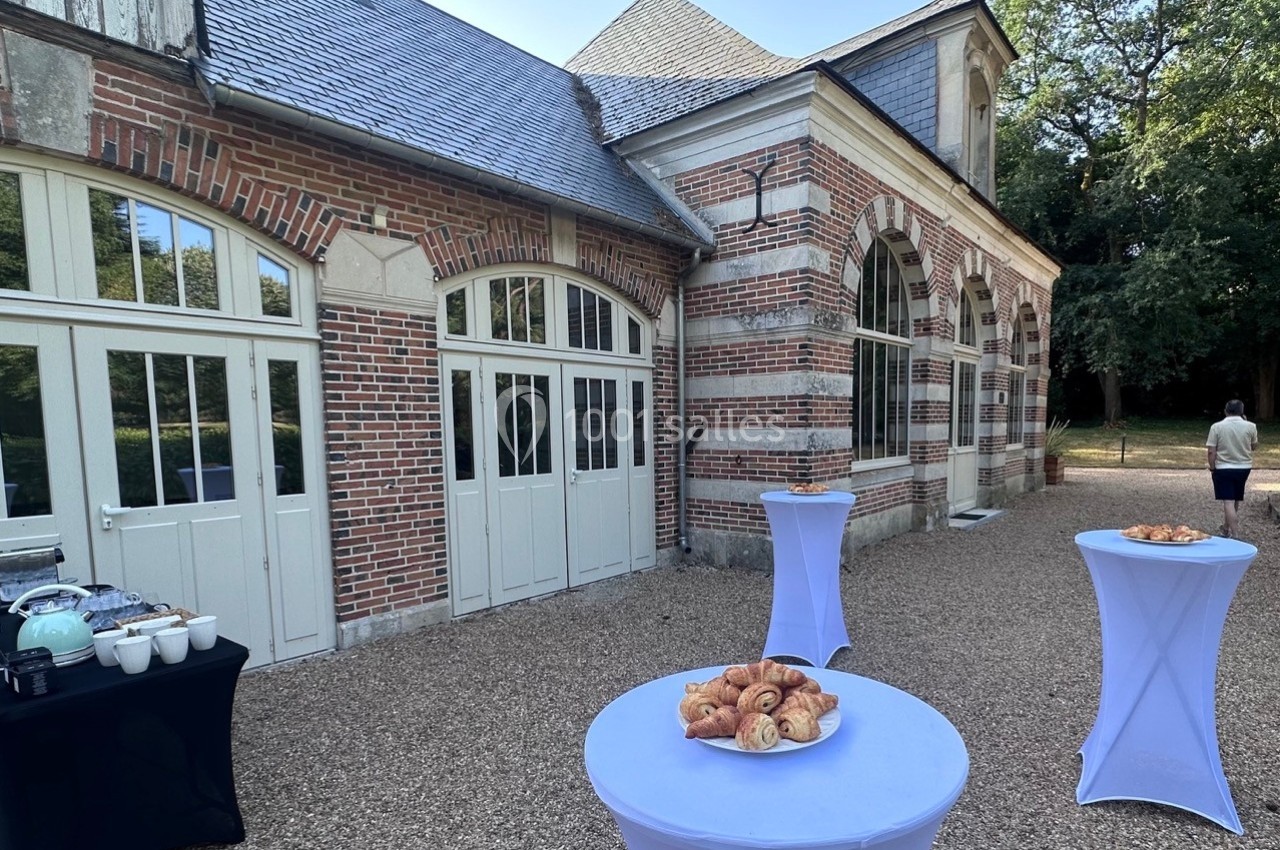 Table dressée avec viennoiseries et boissons devant un bâtiment en briques et grandes fenêtres, dans une cour gravillonnée.