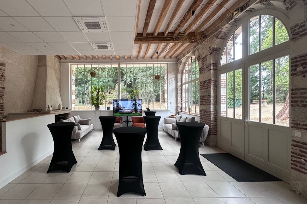 Salle lumineuse avec grandes baies vitrées, tables hautes noires, canapés et écran affichant une image.